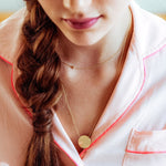 A close-up of a woman wearing a pink shirt and two gold necklaces - a short one with a letter A charm and a long one with a gold solid circle. 