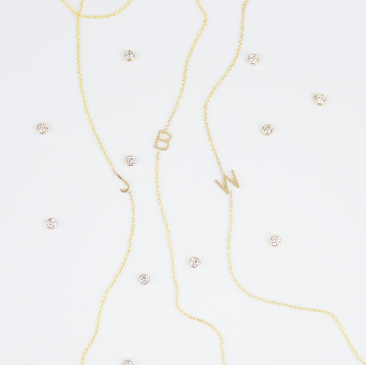 Three gold necklaces with single letter charms, being J, B and W, featuring multiple little gemstones on a white background.
