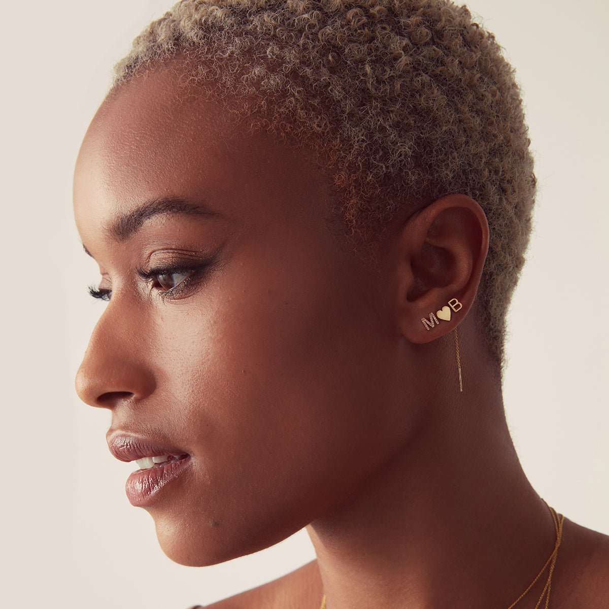 Woman wearing a combination of gold stud earrings - a pavé letter 'M', a heart, and a letter 'B'. 