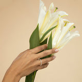 A hand wearing The Lily Boutique Ring holding a bouquet of lilies against a beige background. 