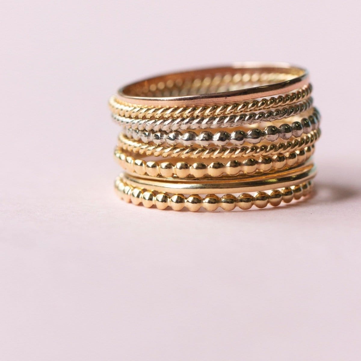 Several Delicate Trio Stacking Rings in yellow gold and white gold on a light pink background. 