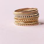Several Delicate Trio Stacking Rings in yellow gold and white gold on a light pink background. 