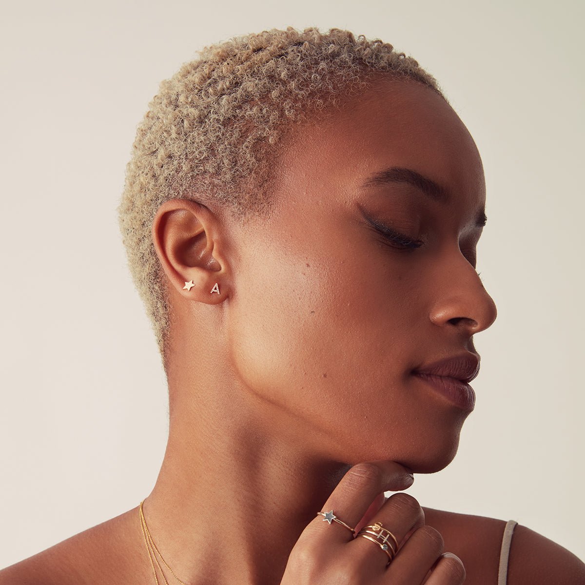 A woman wearing a Heartfelt Stud, a 14K Gold Letter Stud, a 14K Gold Pave Letter Stud, Twinkle Twinkle Ring and a 14K Gold Letter Ring, all in yellow gold, against a light grey background.   