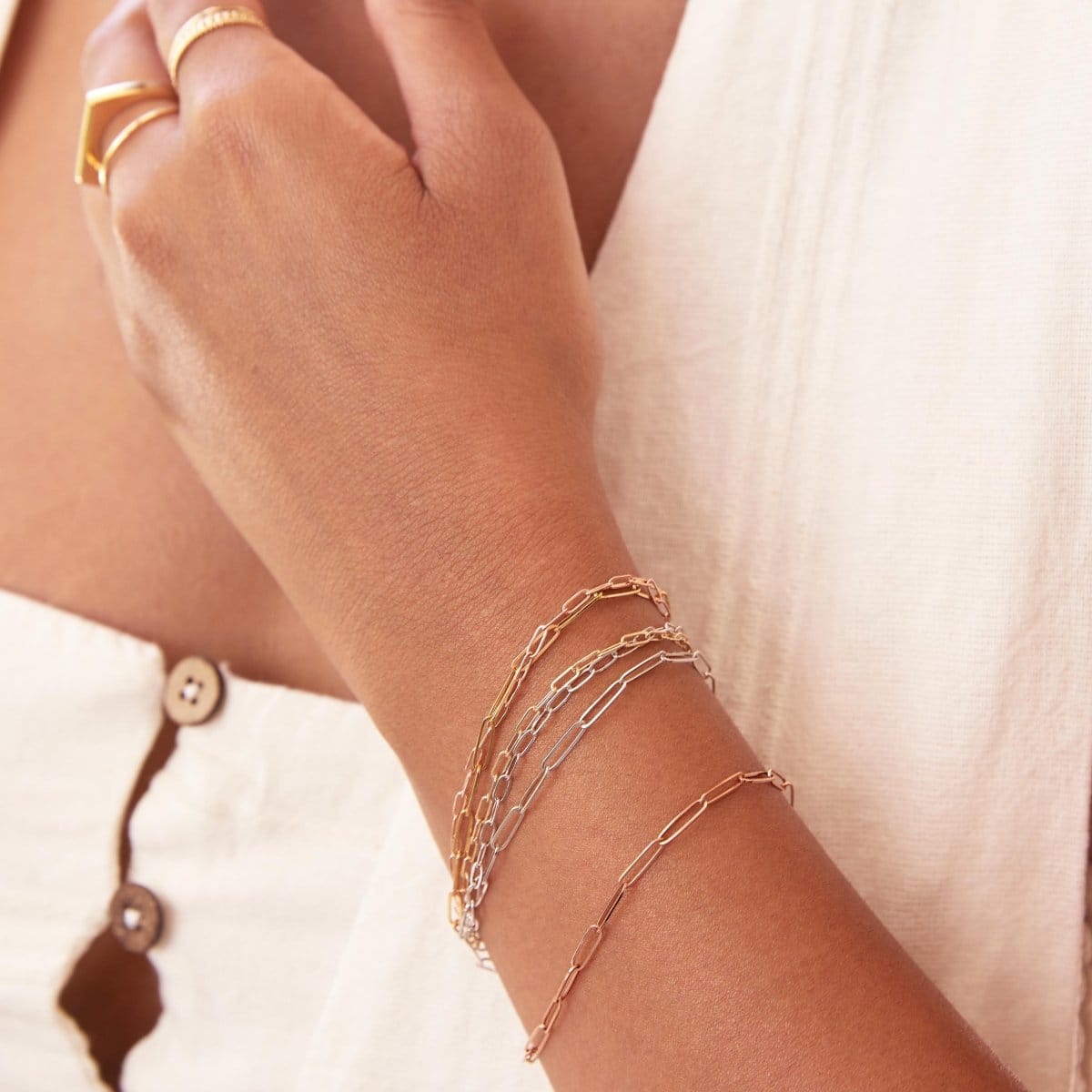 A close-up of a woman's wrist and hand wearing mixed metal Element Short Link Bracelets and Element Long Link Bracelets, and gold rings, styled with a white top. 