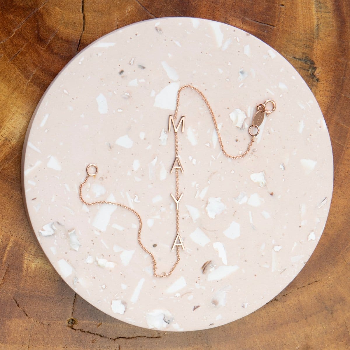A rose gold Custom Gold Bracelet – 4 Letters (M, A, Y, A) laid on a light pink marble round surface against a brown wooden background.