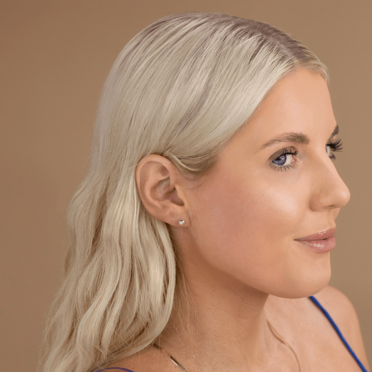 A woman wearing a white gold Poppy Post  with blonde hair against a light brown background. 