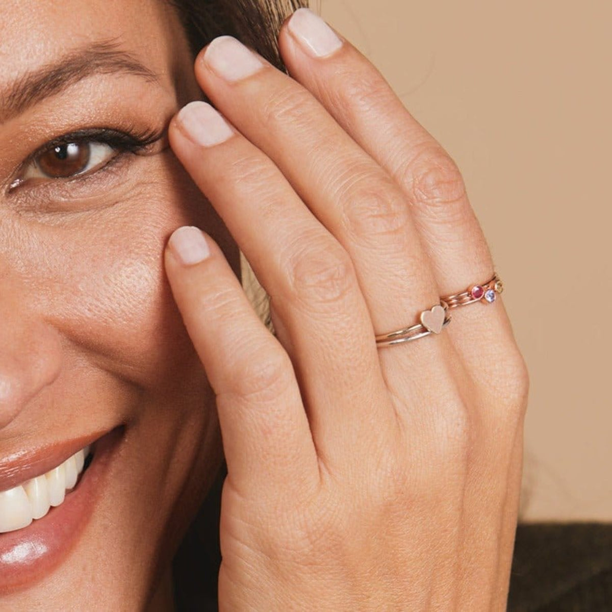 Close-up of a woman's hand wearing a white gold Heartfelt Ring and three Birthstone Rings, against light neutral background. 