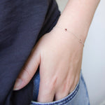 A close-up of a hand wearing a white gold Custom Gold Bracelet - 2 Letters, with a C and a ruby birthstone in a jean pocket, styled with a navy blue top. 