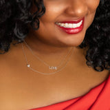 A close-up of a woman's neck wearing a white gold 14k Gold Asymmetrical Charm Necklace - Pavé Heart and a Pavé LOVE Necklace, with black curly hair styled with a red top and lipstick. 