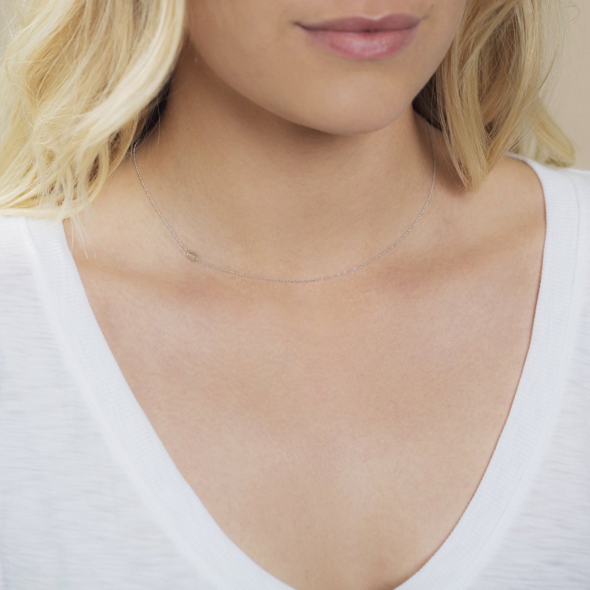 A close-up of a woman's neck wearing a 14k Gold Asymmetrical Number Necklace - 0 in white gold, with blonde hair, styled in a white top.