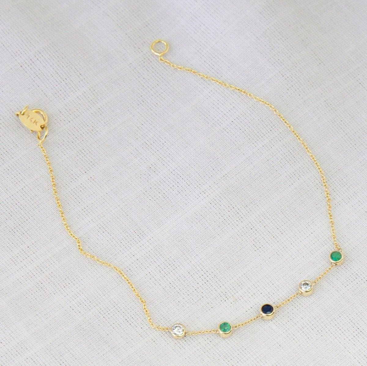 A Custom Gold Bracelet – 5 Letters with five birthstones (two diamond, one sapphire and two emerald) on a white fabric background