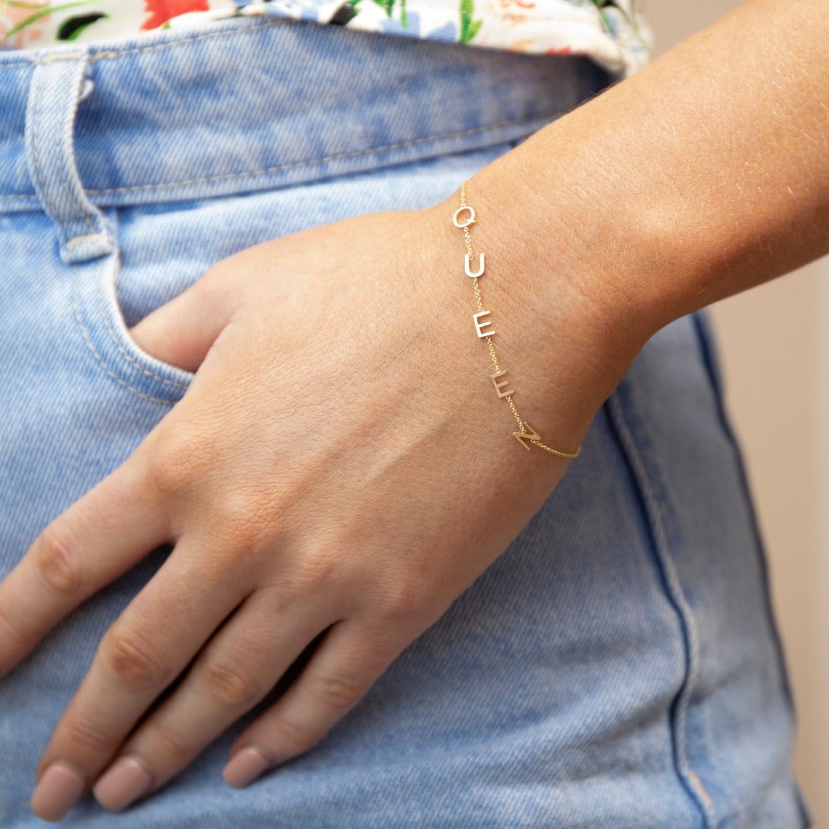 A close-up of a hand wearing a Custom Gold Bracelet – 5 Letters – with the letters Q, U, E, E and N, resting on a blue jean pocket.