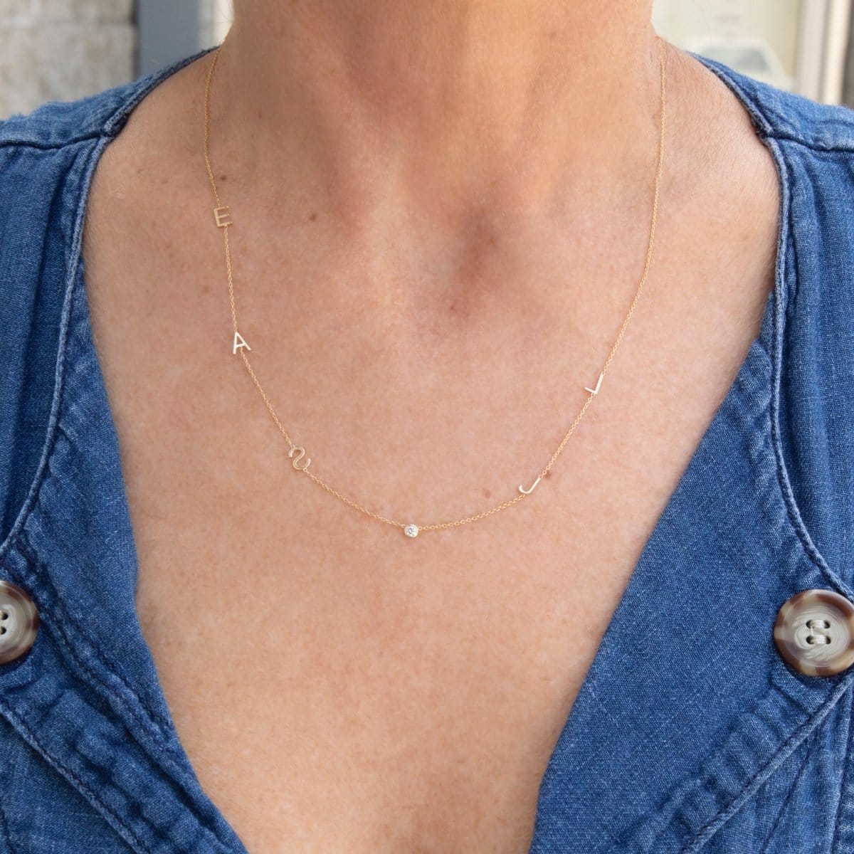 A close-up of a woman’s neck wearing a Custom Gold Necklace in yellow gold with five letter charms (E, A, S, J and L) and a birthstone, in a blue denim shirt.