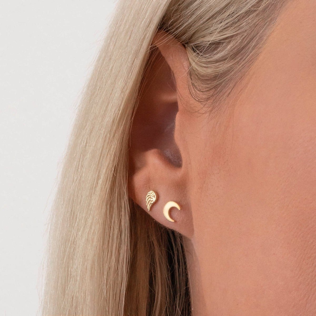 Close-up of Charm Studs - Angel Wing and Moon in yellow gold in a blonde woman's ear against a light grey background. 