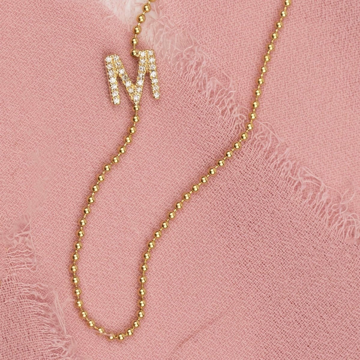 A yellow gold beaded necklace with a pave letter M charm on a pink fabric background. 