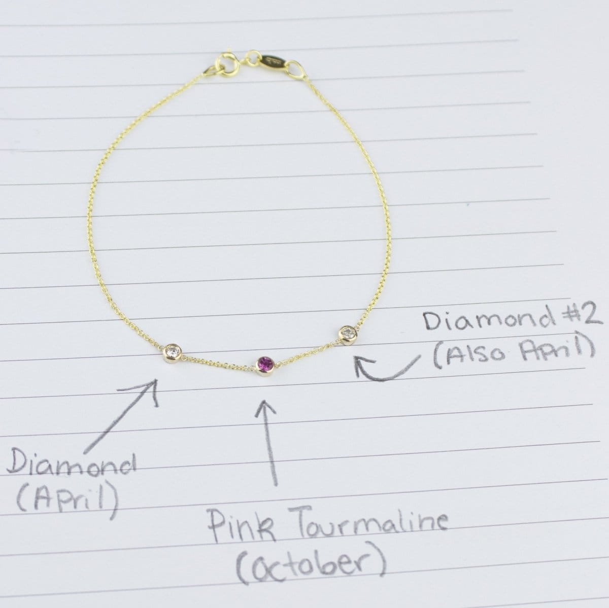 A yellow gold Custom Gold Bracelet with 3 letters and three birthstones (two diamonds and a pink tourmaline) displayed on a sheet of paper with annotations listing the birthstones and their corresponding months.