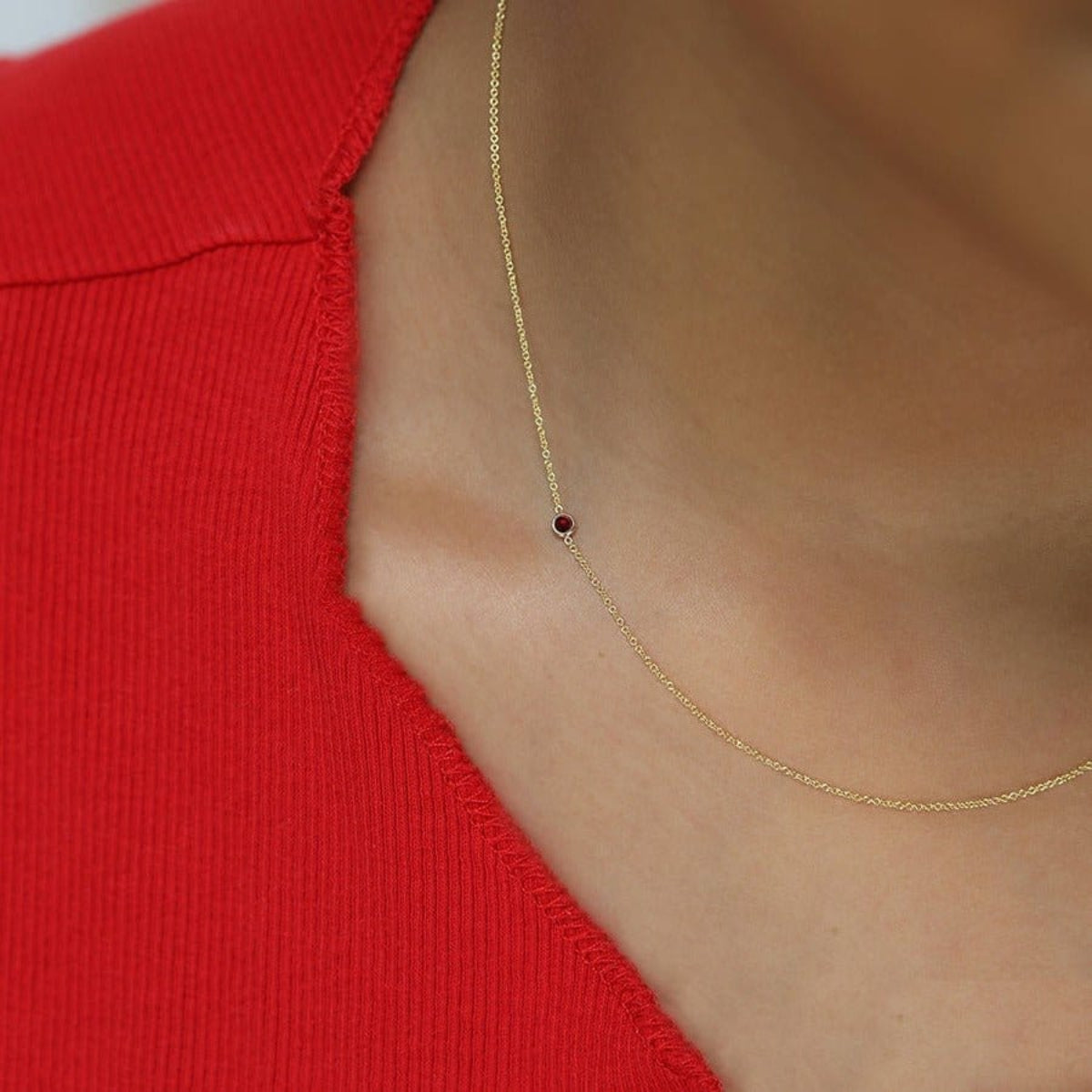 A close-up of a woman's neck wearing a 14K Gold Asymmetrical Birthstone Necklace - Garnet (January) in yellow gold, styled in a red top.