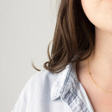 Close-up of a woman's neck and shoulder wearing a 14K Gold Asymmetrical Birthstone Necklace - Peridot (August) in yellow gold, with brown hair, styled in a light blue denim shirt against a light grey background.