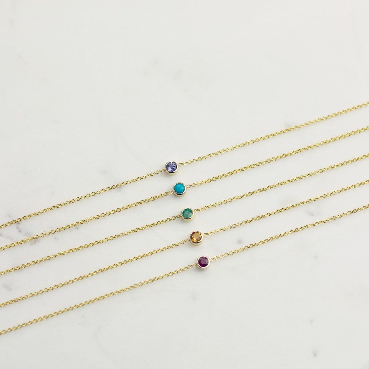 Five yellow gold 14K Gold Asymmetrical Birthstone Necklaces - Tanzanite (December), Turquoise (December), Emerald (May), Citrine (November) and Garnet (January) laid on a white background. 