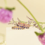 The Peony Bouquet Ring, The Violet Bouquet Ring and The Periwinkle Bouquet Ring against a light yellow blurred background with purple flowers. 