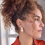 Close-up of a Cascading Solid Eclipse Earring in yellow gold being worn by a woman with brown curly hair and a red top against a light blurred background. 