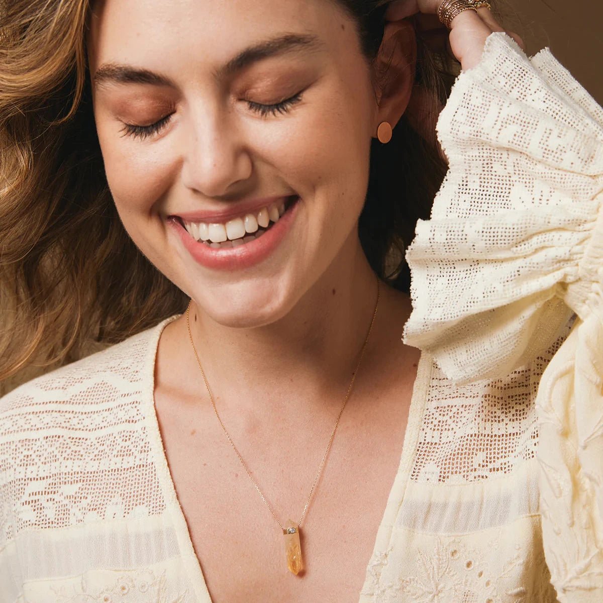 A woman wearing The Happiness Retreat Necklace, a Solid Eclipse Earring - Small and several rings, styled in a light yellow lace top with a warm background.