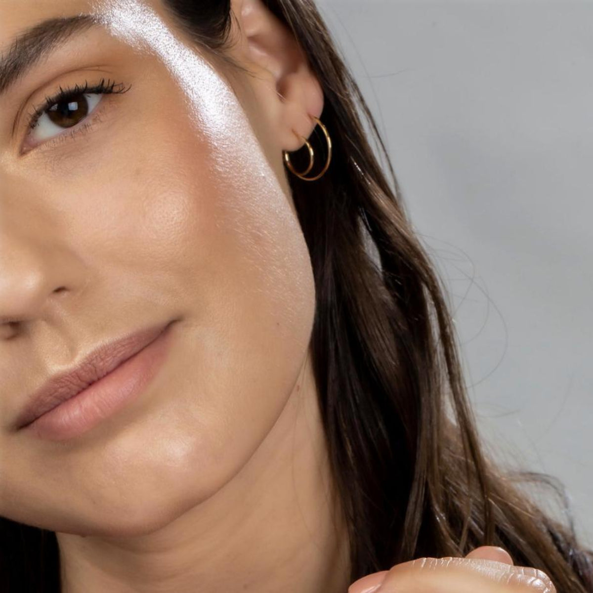 Close-up of a brunette woman wearing  yellow gold Crescent Hoops (one Small and Medium) against a light grey background. 