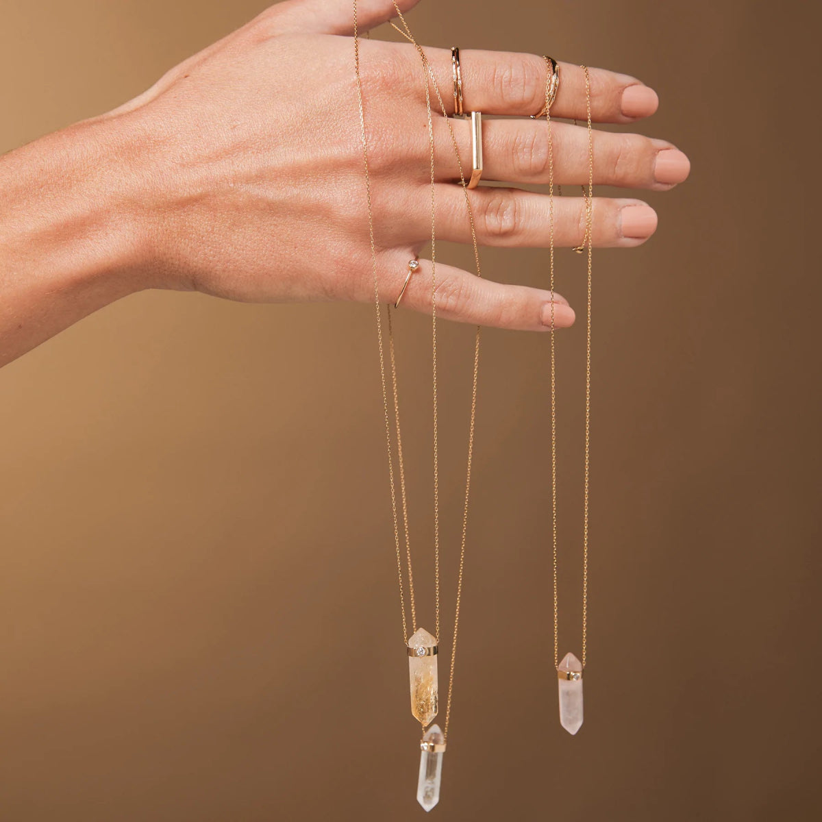 A hand holding three Retreat Necklaces in Clarity, Happiness and Love, and wearing 4 Diamond Rings, a Geo Stacking Ring Square and a Birthstone Ring against a light brown background. 