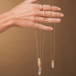 A hand holding three Retreat Necklaces in Clarity, Happiness and Love, and wearing 4 Diamond Rings, a Geo Stacking Ring Square and a Birthstone Ring against a light brown background. 