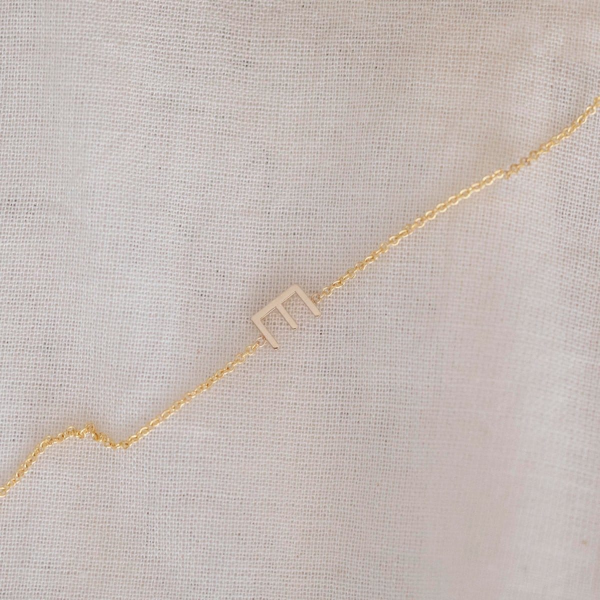 A yellow gold necklace with a letter E charm, displayed diagonally on a cream fabric background.