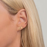 Close-up of a Charm Stud - Hamsa and Charm Stud - Star of David in yellow gold in a blonde woman's ear against a light grey background. 