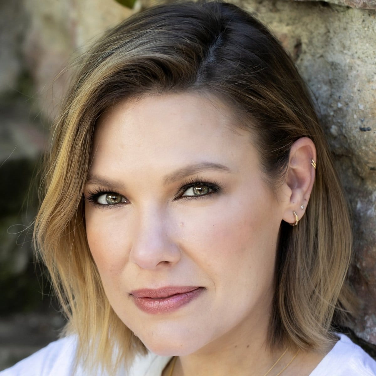 A woman wearing an Alli Webb Faceted Huggie, a stud earring and two Rounded Ear Cuffs in yellow gold, with short hair against a stone background.