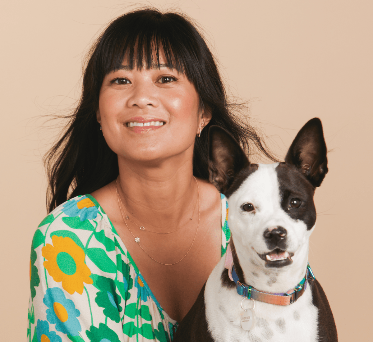 A woman wearing a yellow gold 14k Gold Asymmetrical Charm Necklace - Paw and a Custom Gold Necklace - 2 Letters, wearing a green, blue and yellow floral top, holding a dog against a beige background.