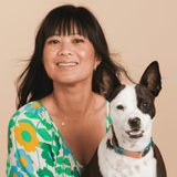 A woman wearing a yellow gold 14k Gold Asymmetrical Charm Necklace - Paw and a Custom Gold Necklace - 2 Letters, wearing a green, blue and yellow floral top, holding a dog against a beige background.