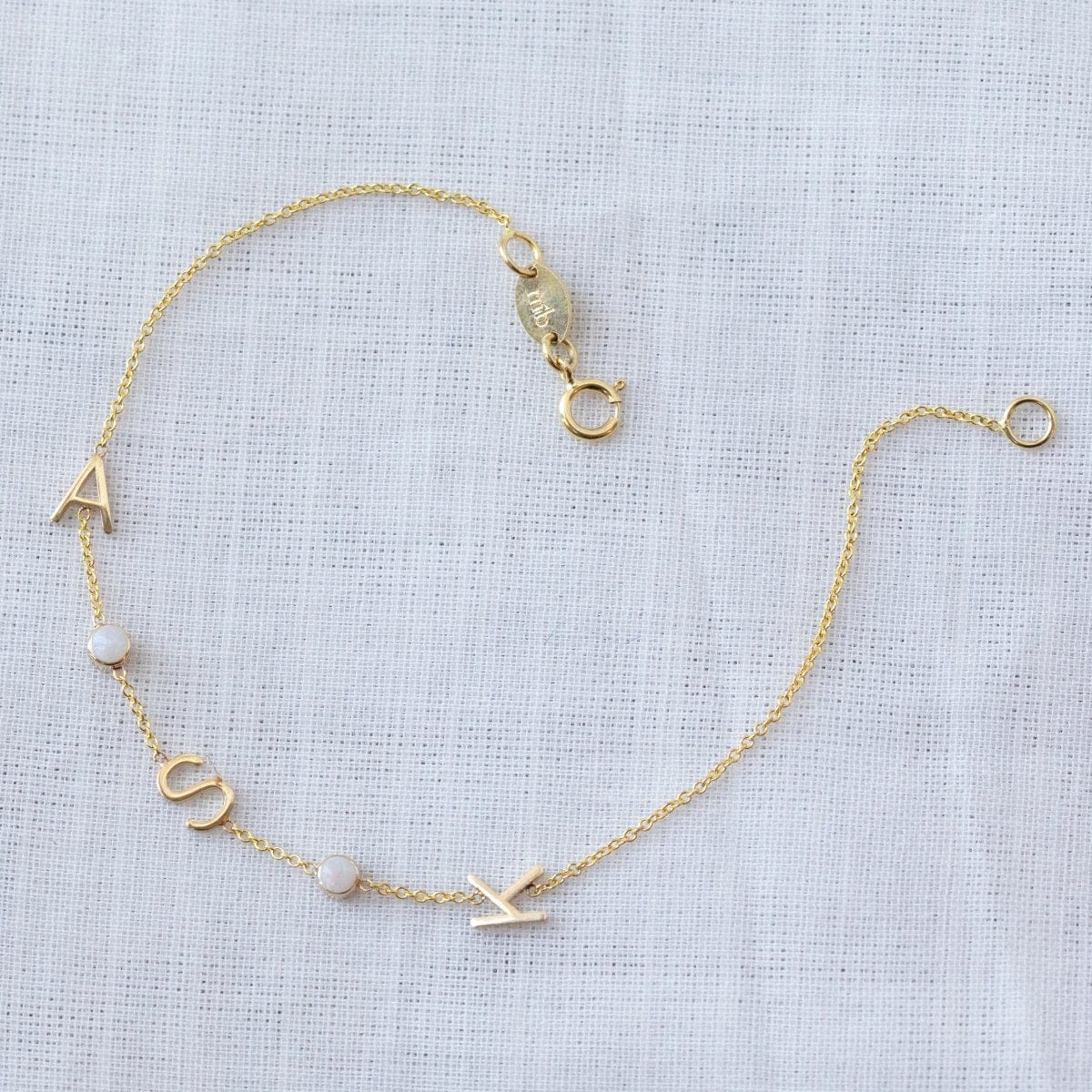 A yellow gold Custom Gold Bracelet - 5 letters, with the letter charms A, S and K and two birthstones on a white fabric background. 