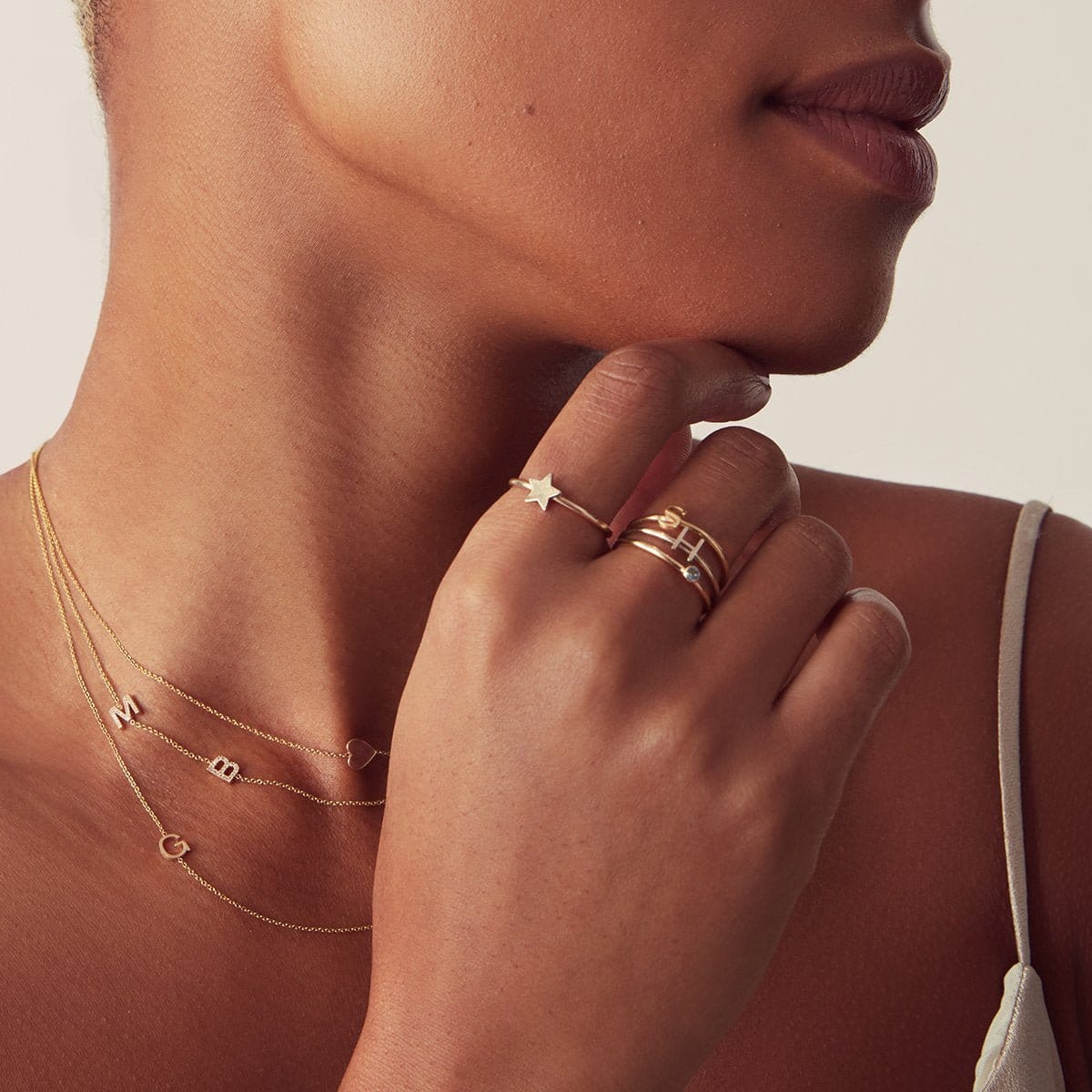 Close-up of a woman's hand and neck wearing multiple gold letter and charm necklaces and rings against a neutral background. 