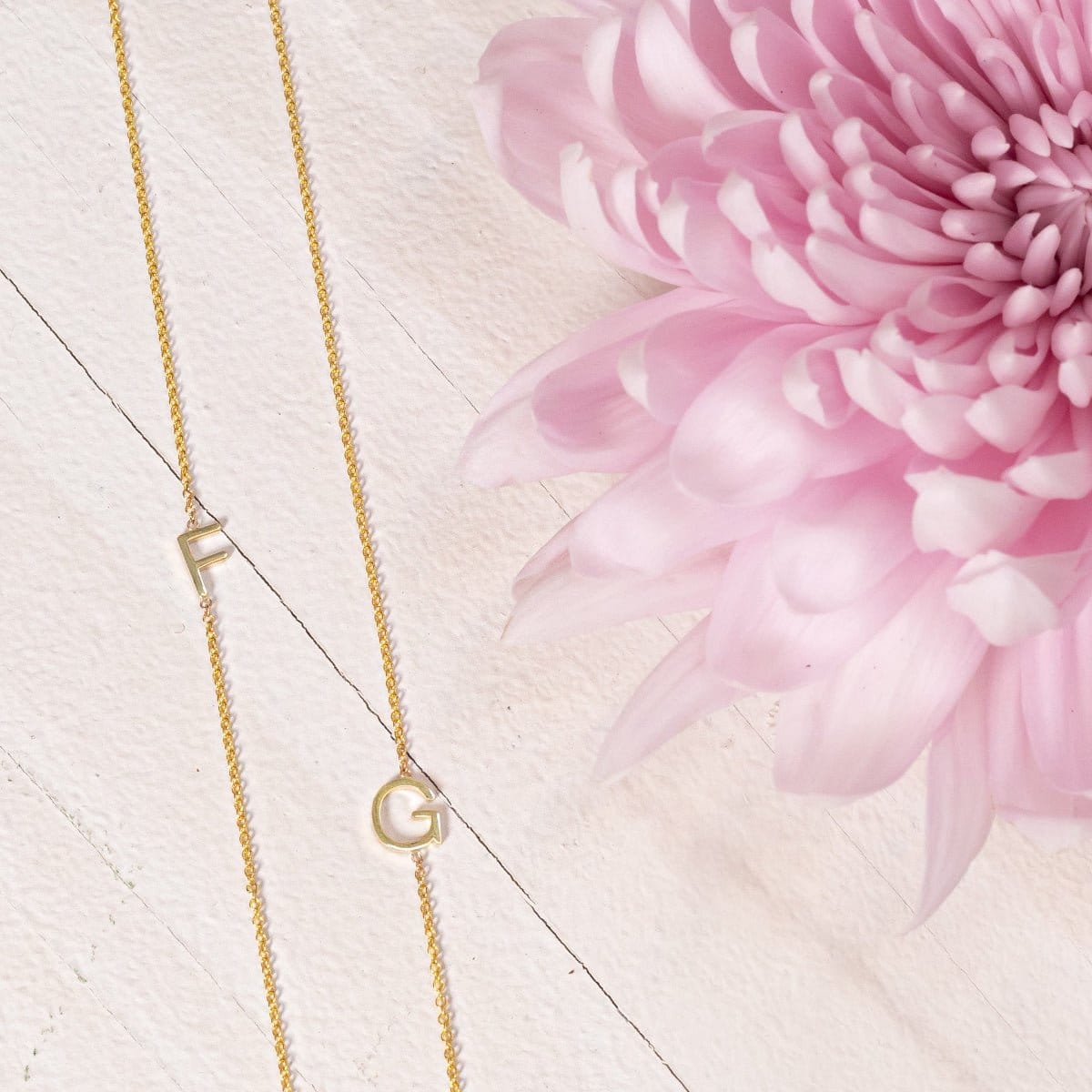 Two yellow gold necklaces with single letter charms, F and G, displayed on a neutral background with a pink flower.
