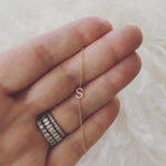 A close-up of a hand wearing three rings, with a yellow gold necklace featuring a letter S charm wrapped around the fingers, set against a cream furry background.