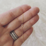 A close-up of a hand wearing three rings, with a yellow gold necklace featuring a letter S charm wrapped around the fingers, set against a cream furry background.