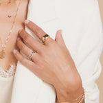 Close-up of a woman’s hand wearing a Link Ring, two Pavé Diamond Rings, a Paris Ring, an Off the Cuff Bracelet, and a Bubble Bracelet, styled with a 14k gold Asymmetrical Charm Necklace (Moon) and other yellow gold necklaces, paired with a white blazer and lace top.