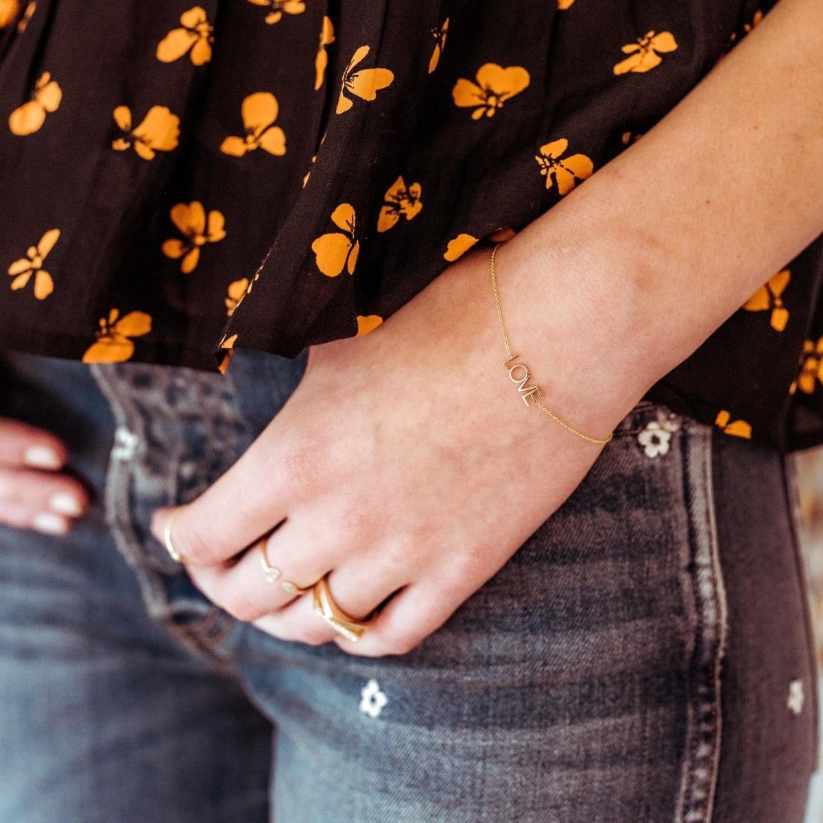 Close-up of a person arm and hand wearing a Love Bracelet and gold rings, including a Geo Stacking Ring, Pave Geo Ring with a black and yellow floral-patterned shirt and blue jeans. 
