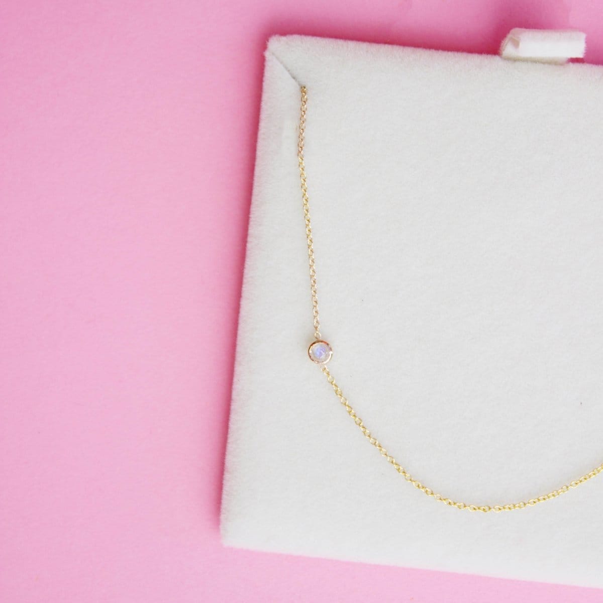 A 14K Gold Asymmetrical Birthstone Necklace - Moonstone (June) in a white cushion case, against a pink background. 