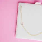 A 14K Gold Asymmetrical Birthstone Necklace - Moonstone (June) in a white cushion case, against a pink background. 