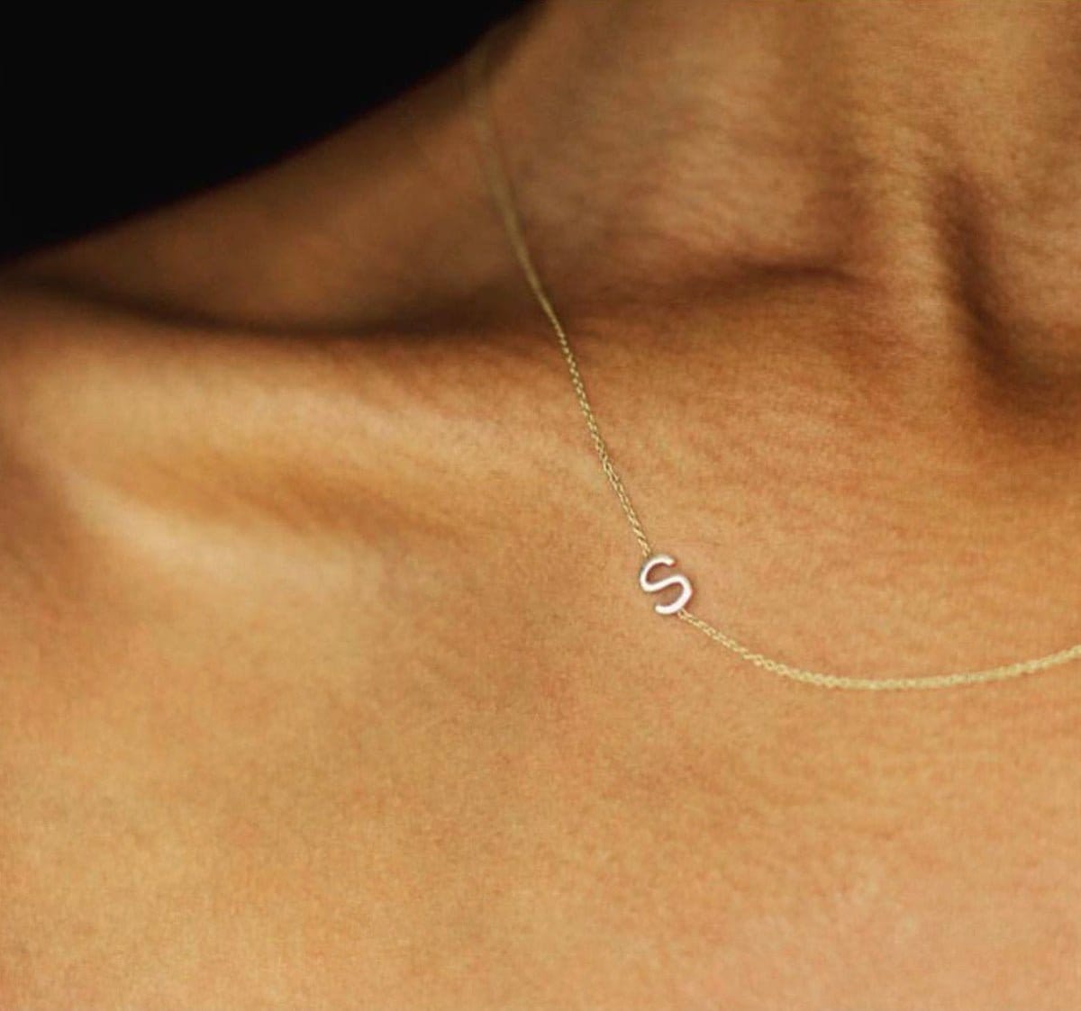 A close up of a woman's décolletage wearing a yellow gold necklace with a letter S charm. 