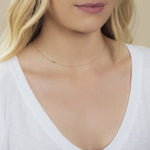 A close-up of a woman's neck wearing a 14k Gold Asymmetrical Number Necklace - 7 in yellow gold, styled with blonde hair and a white top.