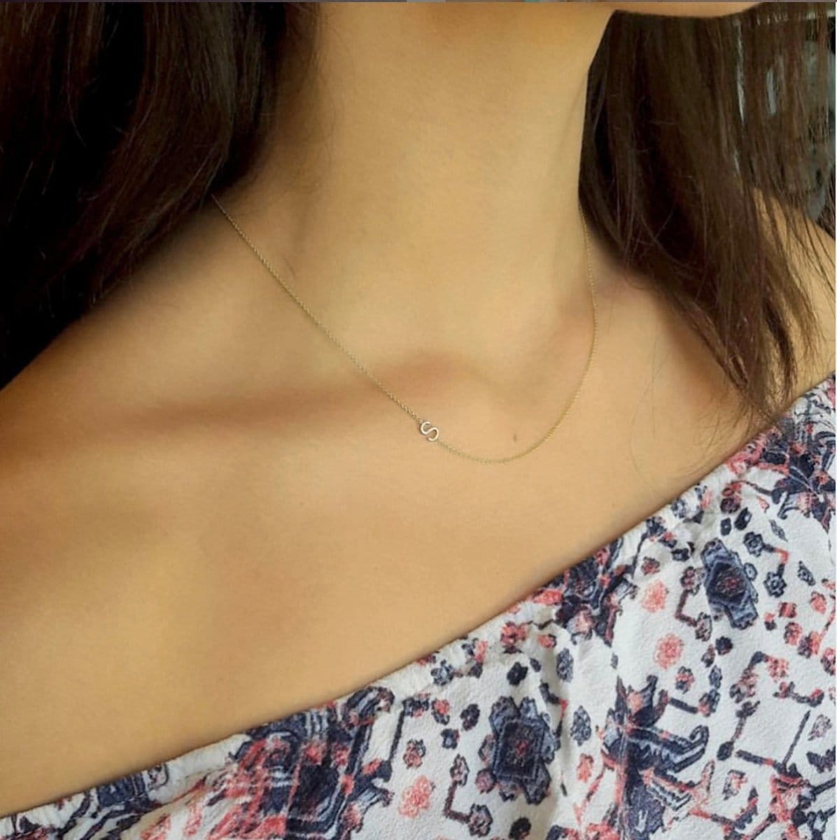 A close-up of a woman’s neck area wearing a yellow gold necklace with a letter S charm and a patterned off-shoulder top.