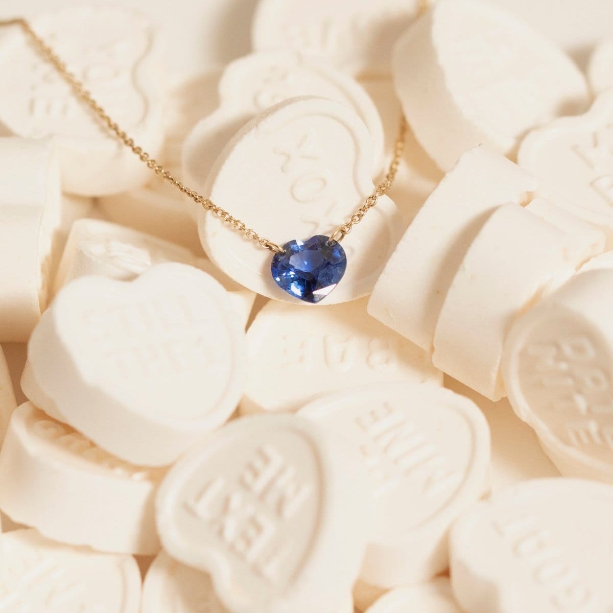 The Sweetheart Necklace - Blue Sapphire in yellow gold laid on top of white heart-shaped candy. 