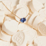 The Sweetheart Necklace - Blue Sapphire in yellow gold laid on top of white heart-shaped candy. 