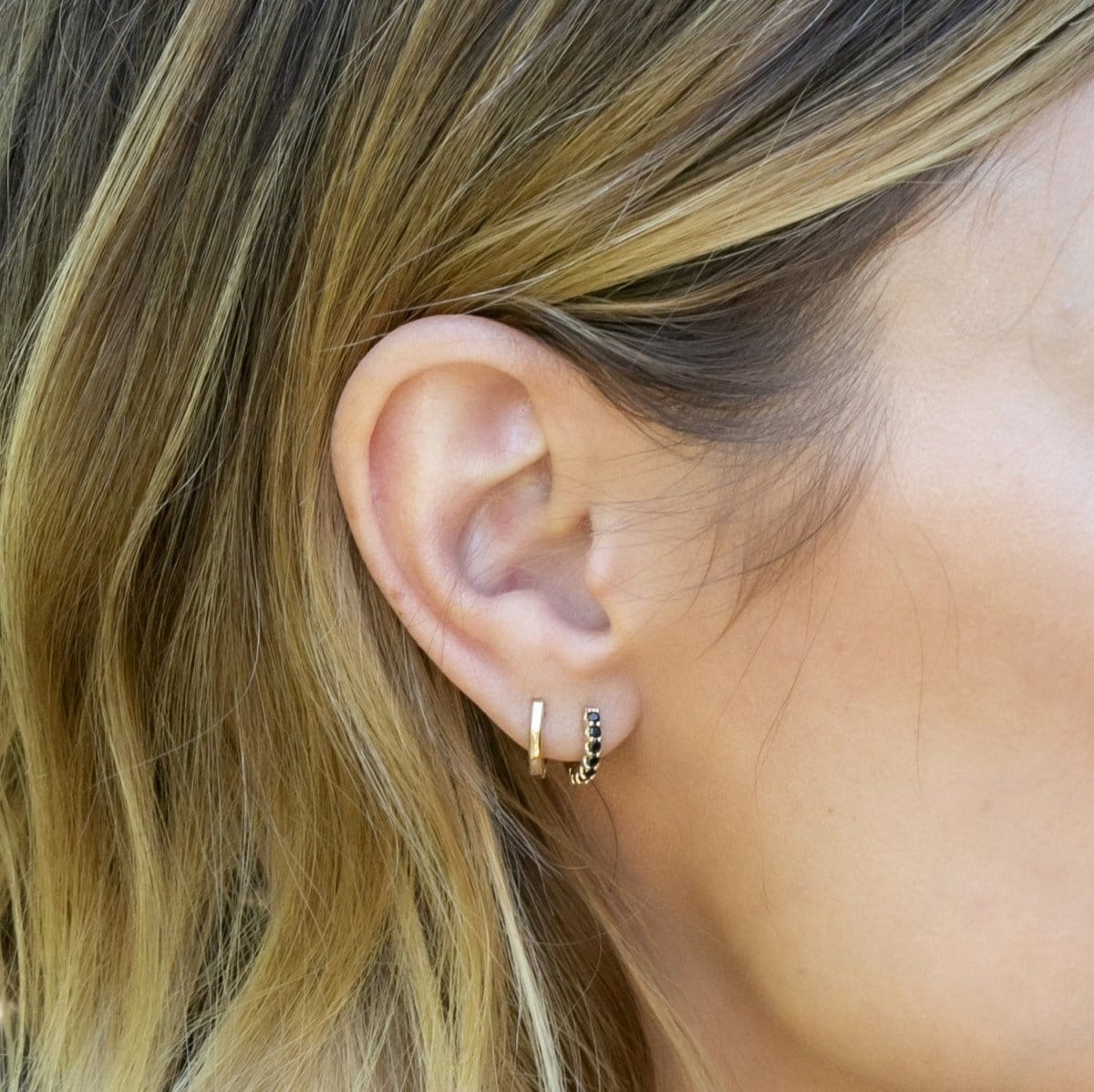 Close-up of a woman's ear wearing an Alli Webb Faceted Huggie and Onyx Huggie, with her blonde hair visible. 
