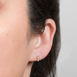 Close-up of an ear wearing a Pave Charm Stud - Cross in yellow gold against a light grey background.